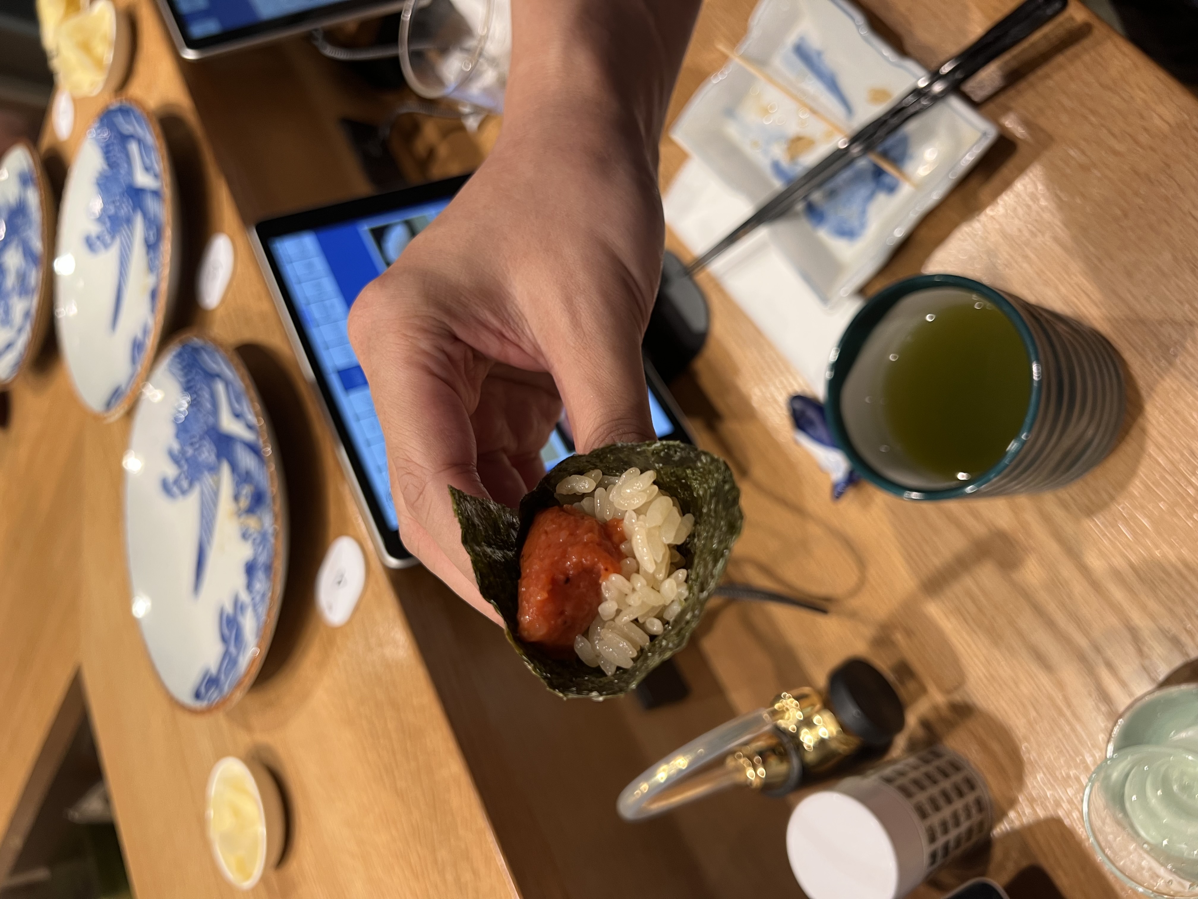 Temaki hand roll being held, with sake and ceramic plates visible on the counter